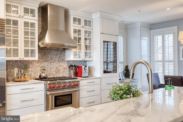 kitchen with tasteful backsplash, white cabinets, high end stainless steel range oven, light stone countertops, and wall chimney exhaust hood