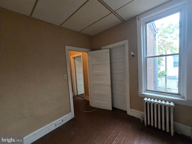 unfurnished bedroom featuring a paneled ceiling, radiator heating unit, dark wood-type flooring, and multiple windows