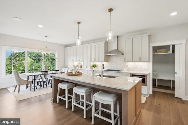 kitchen featuring light stone counters, stainless steel oven, a center island with sink, wall chimney range hood, and white cabinetry