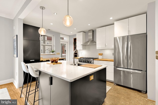 kitchen featuring wall chimney exhaust hood, decorative light fixtures, white cabinets, an island with sink, and stainless steel refrigerator