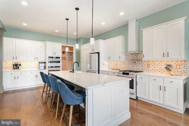 kitchen with wall chimney exhaust hood, high end white fridge, gas stove, white cabinets, and an island with sink