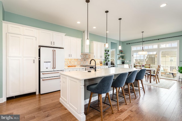 kitchen featuring pendant lighting, white refrigerator with ice dispenser, a center island with sink, white cabinets, and wall chimney range hood