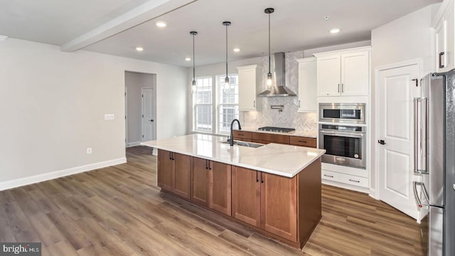 kitchen featuring white cabinetry, wall chimney exhaust hood, stainless steel appliances, light stone counters, and a kitchen island with sink