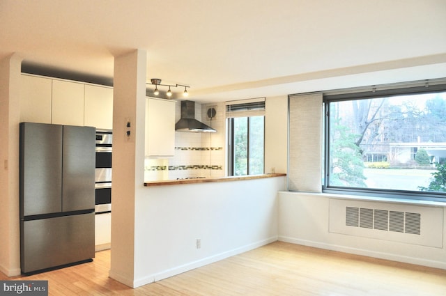 kitchen featuring radiator, a wealth of natural light, wall chimney range hood, white cabinets, and appliances with stainless steel finishes