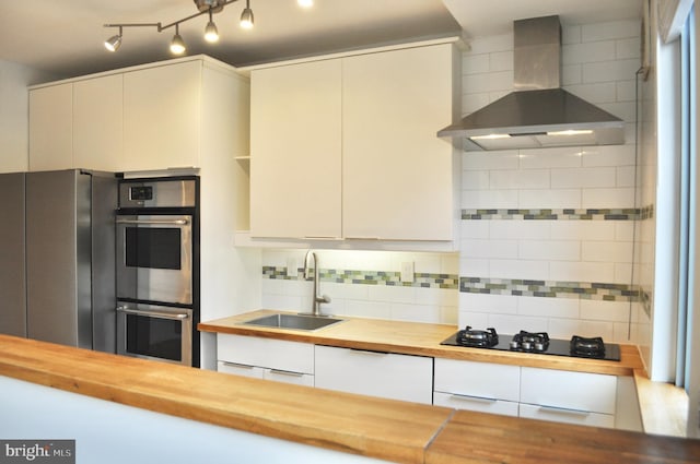 kitchen with wooden counters, wall chimney exhaust hood, appliances with stainless steel finishes, tasteful backsplash, and white cabinetry