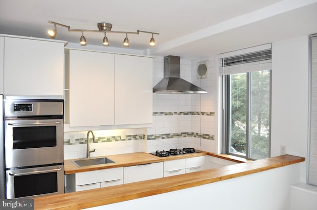 kitchen featuring wooden counters, sink, wall chimney range hood, black gas cooktop, and white cabinetry