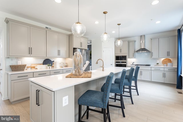 kitchen featuring appliances with stainless steel finishes, an island with sink, gray cabinets, and wall chimney range hood