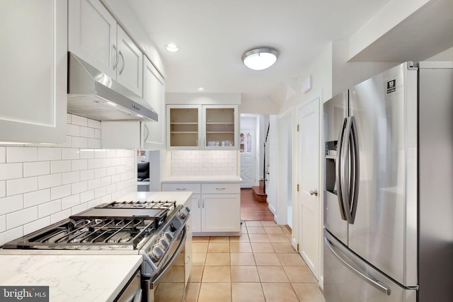 kitchen featuring white cabinets, decorative backsplash, range hood, light tile patterned flooring, and stainless steel appliances