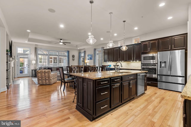 kitchen with light stone countertops, appliances with stainless steel finishes, a tray ceiling, decorative light fixtures, and a center island with sink