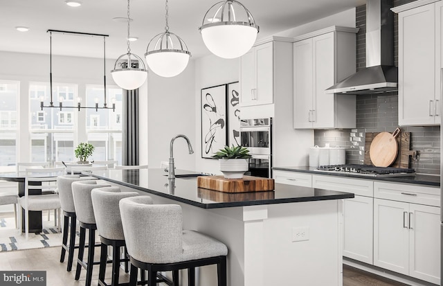 kitchen with a center island with sink, wall chimney range hood, hanging light fixtures, and appliances with stainless steel finishes