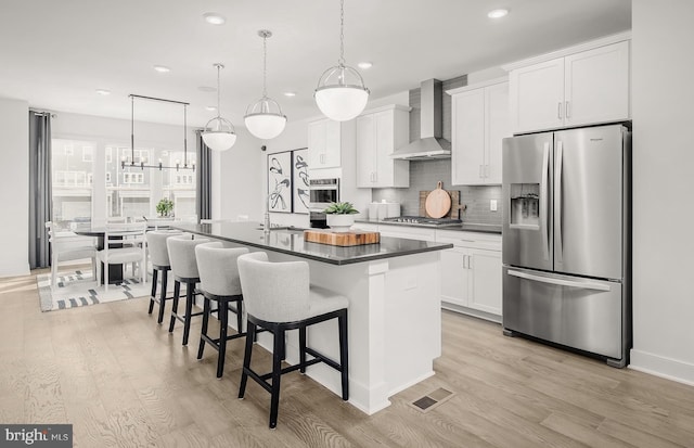 kitchen with wall chimney exhaust hood, an island with sink, light hardwood / wood-style floors, white cabinets, and appliances with stainless steel finishes