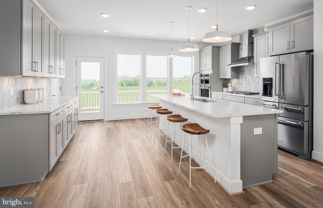 kitchen featuring appliances with stainless steel finishes, light wood-type flooring, gray cabinetry, wall chimney range hood, and a center island with sink