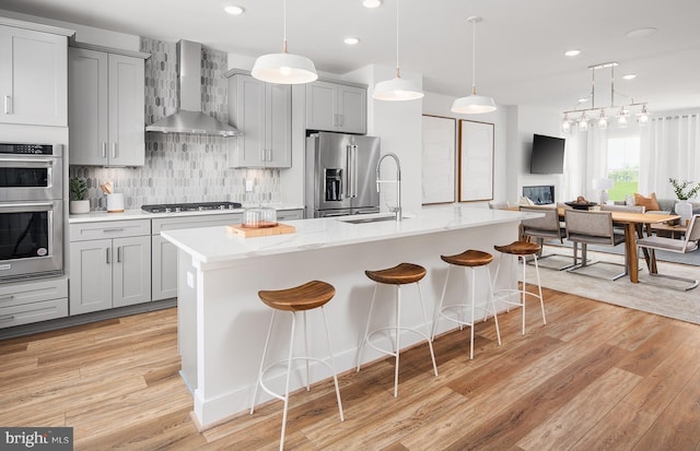 kitchen featuring stainless steel appliances, wall chimney range hood, a center island with sink, light hardwood / wood-style flooring, and gray cabinets