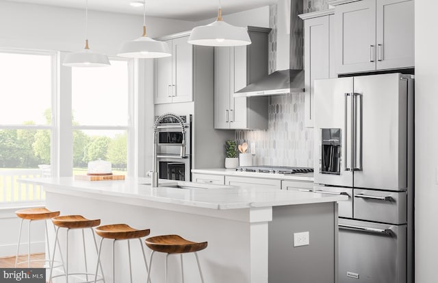 kitchen with light stone counters, stainless steel appliances, sink, wall chimney range hood, and a center island with sink