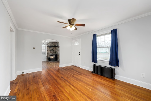 unfurnished living room featuring a wood stove, radiator, ceiling fan with notable chandelier, hardwood / wood-style flooring, and ornamental molding