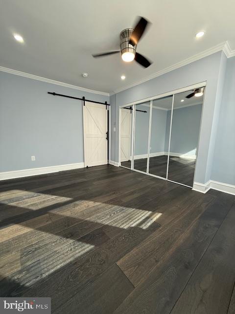 unfurnished bedroom with dark hardwood / wood-style flooring, ceiling fan, crown molding, a barn door, and a closet