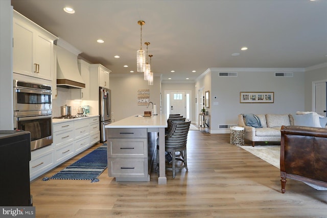 kitchen with stainless steel appliances, pendant lighting, a center island with sink, white cabinets, and custom range hood