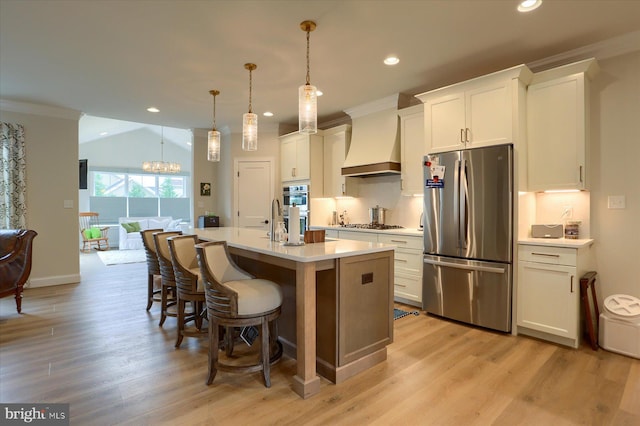 kitchen featuring appliances with stainless steel finishes, premium range hood, decorative light fixtures, white cabinetry, and an island with sink