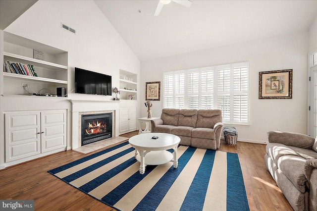 living room featuring built in shelves, ceiling fan, wood-type flooring, high vaulted ceiling, and a tiled fireplace