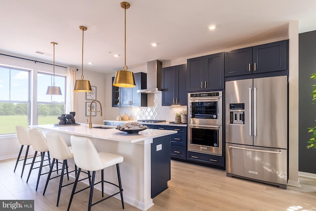 kitchen with appliances with stainless steel finishes, wall chimney exhaust hood, decorative light fixtures, a breakfast bar area, and an island with sink