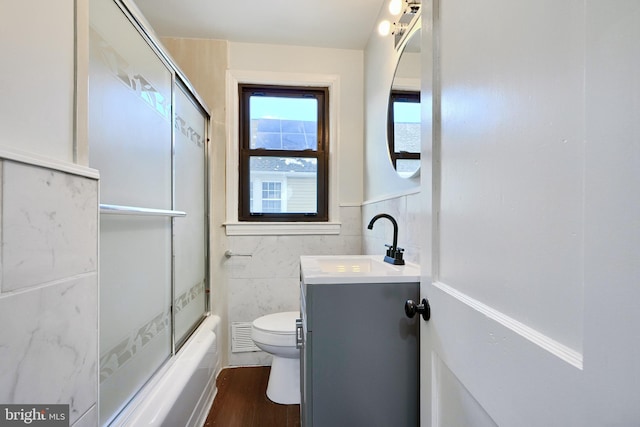 full bathroom featuring toilet, shower / bath combination with glass door, tile walls, wood-type flooring, and vanity