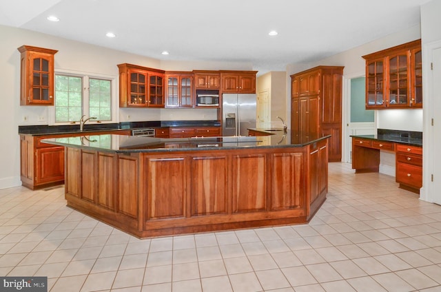 kitchen with dark countertops, a kitchen island with sink, built in study area, stainless steel appliances, and glass insert cabinets