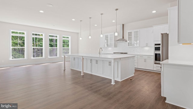 kitchen featuring wall chimney exhaust hood, wood-type flooring, white cabinetry, hanging light fixtures, and an island with sink