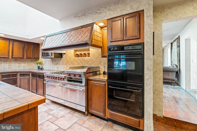 kitchen with high end range, tasteful backsplash, premium range hood, black double oven, and tile counters