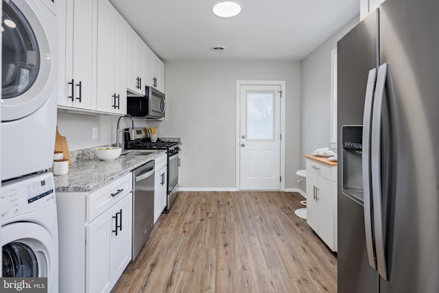 kitchen with light stone countertops, light wood-type flooring, stainless steel appliances, stacked washer / dryer, and white cabinets