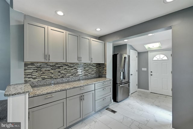 kitchen with gray cabinets, decorative backsplash, stainless steel fridge, and light stone counters