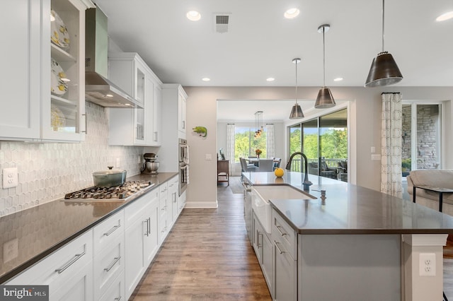 kitchen featuring stainless steel appliances, wall chimney range hood, decorative light fixtures, white cabinets, and an island with sink