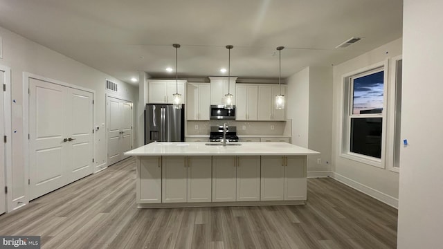 kitchen with appliances with stainless steel finishes, decorative light fixtures, a center island with sink, light hardwood / wood-style floors, and white cabinetry