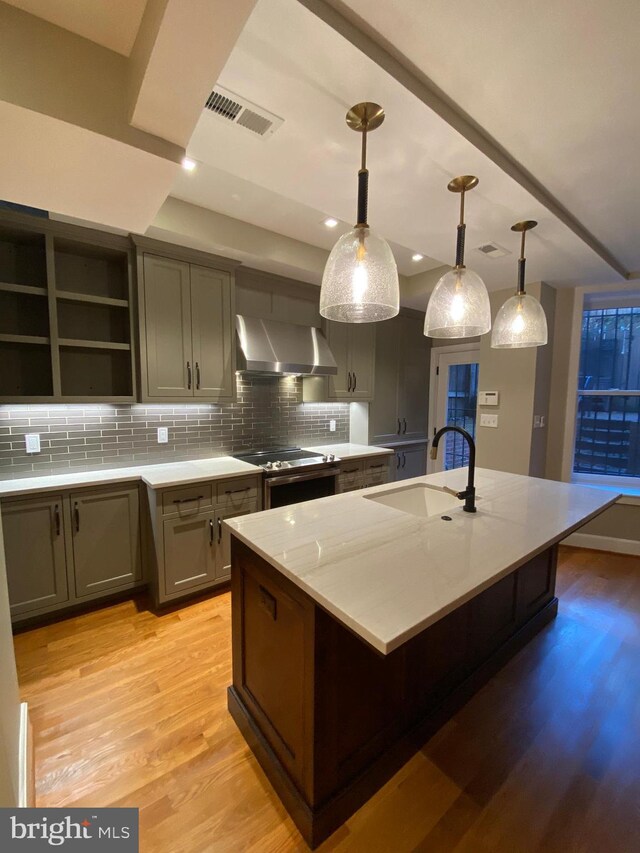 kitchen with light wood-type flooring, wall chimney exhaust hood, sink, pendant lighting, and a center island with sink