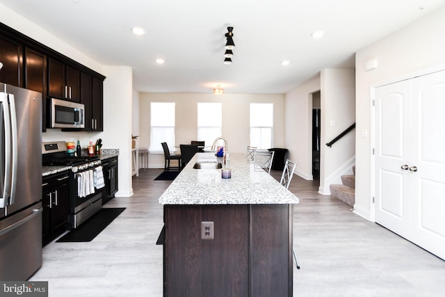 kitchen with light stone countertops, sink, dark brown cabinets, stainless steel appliances, and a center island with sink