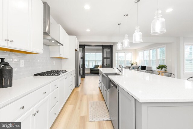 kitchen featuring appliances with stainless steel finishes, wall chimney exhaust hood, decorative light fixtures, white cabinetry, and an island with sink