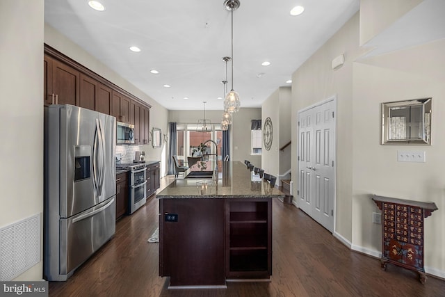 kitchen with dark brown cabinetry, stainless steel appliances, a kitchen island with sink, sink, and hanging light fixtures