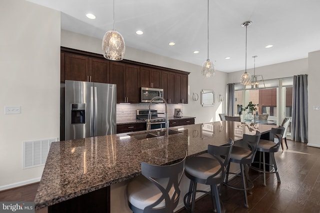 kitchen featuring dark stone counters, stainless steel appliances, hanging light fixtures, and a center island with sink