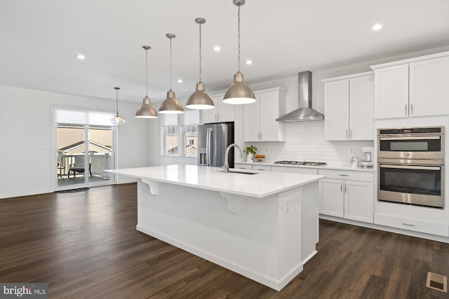 kitchen with stainless steel appliances, wall chimney range hood, pendant lighting, white cabinets, and an island with sink