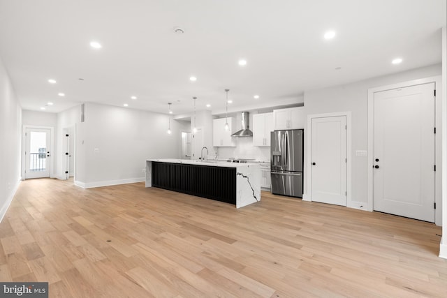 kitchen featuring wall chimney exhaust hood, stainless steel refrigerator with ice dispenser, decorative light fixtures, white cabinetry, and an island with sink