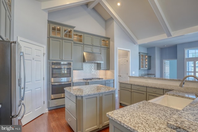 kitchen with appliances with stainless steel finishes, a kitchen island, sink, lofted ceiling with beams, and light stone counters