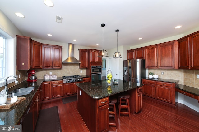 kitchen with sink, stainless steel appliances, wall chimney range hood, decorative light fixtures, and a kitchen island