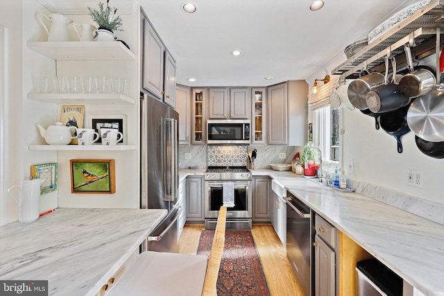 kitchen with gray cabinetry, stainless steel appliances, tasteful backsplash, light stone counters, and light hardwood / wood-style floors