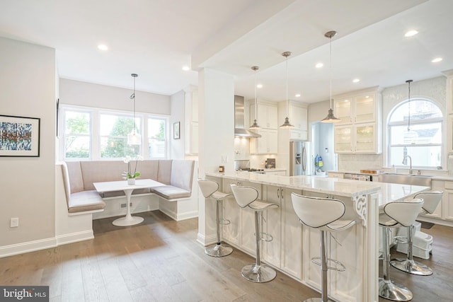 kitchen featuring light stone counters, wall chimney range hood, stainless steel fridge with ice dispenser, white cabinetry, and a breakfast bar area