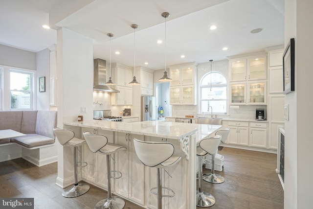 kitchen with stainless steel refrigerator with ice dispenser, backsplash, a kitchen breakfast bar, wall chimney range hood, and white cabinets