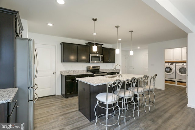 kitchen with washing machine and clothes dryer, sink, hanging light fixtures, stainless steel appliances, and a center island with sink