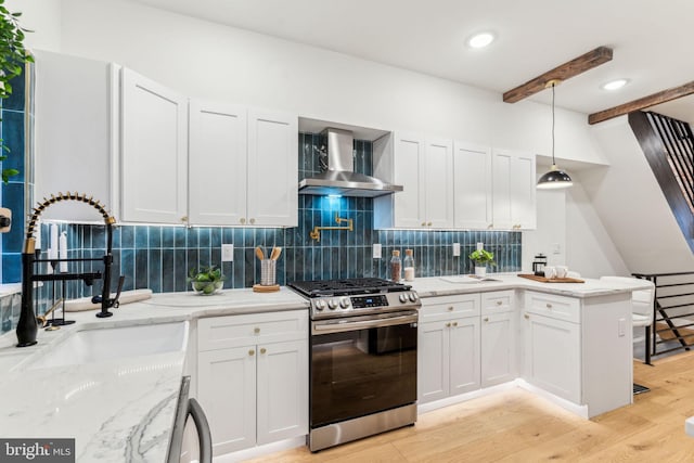 kitchen featuring wall chimney exhaust hood, stainless steel gas range oven, white cabinetry, and hanging light fixtures