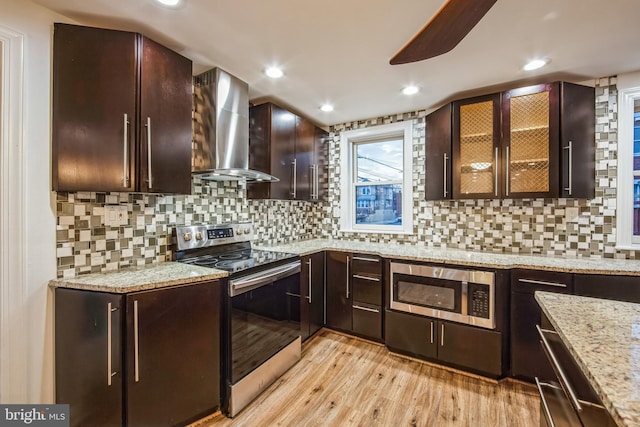 kitchen featuring stainless steel appliances, wall chimney range hood, light stone counters, backsplash, and light hardwood / wood-style floors