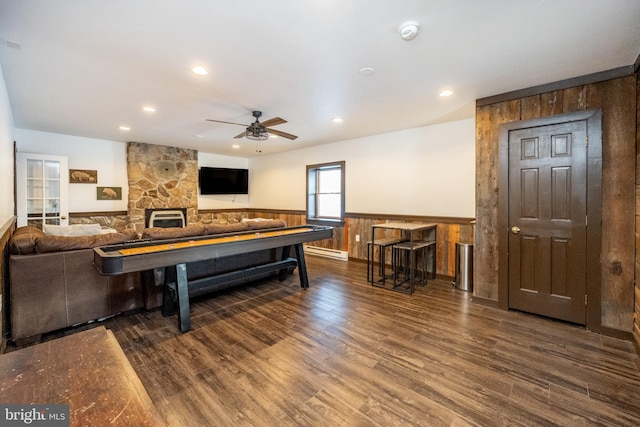 game room with dark hardwood / wood-style flooring, baseboard heating, ceiling fan, a stone fireplace, and wood walls