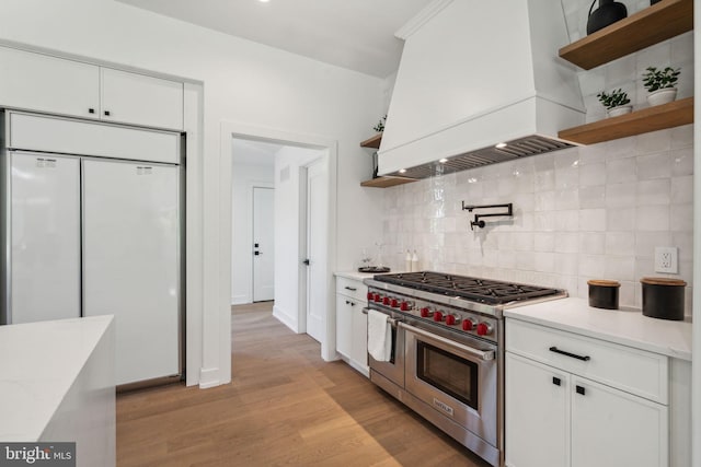 kitchen with decorative backsplash, custom range hood, high end appliances, white cabinets, and light hardwood / wood-style floors