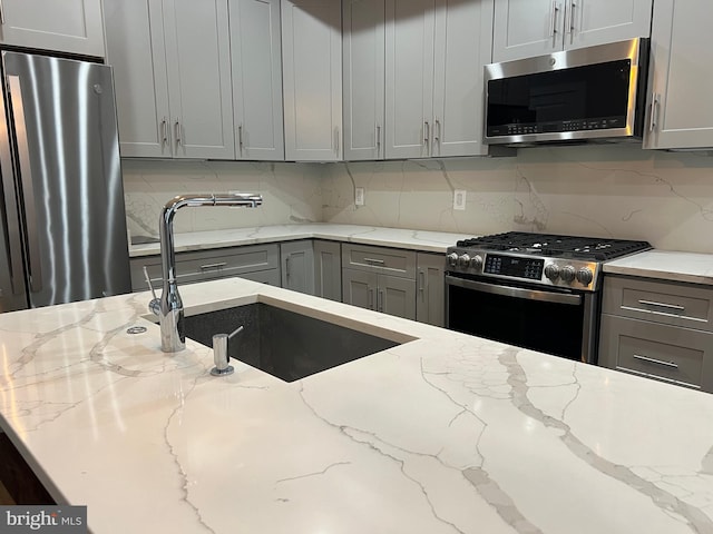 kitchen with gray cabinetry, light stone countertops, sink, stainless steel appliances, and backsplash